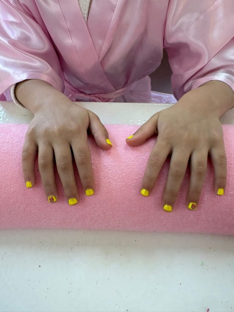 Close-up of two hands resting on a pink surface; nails painted bright yellow, with a small flower design on the left-hand ring finger.