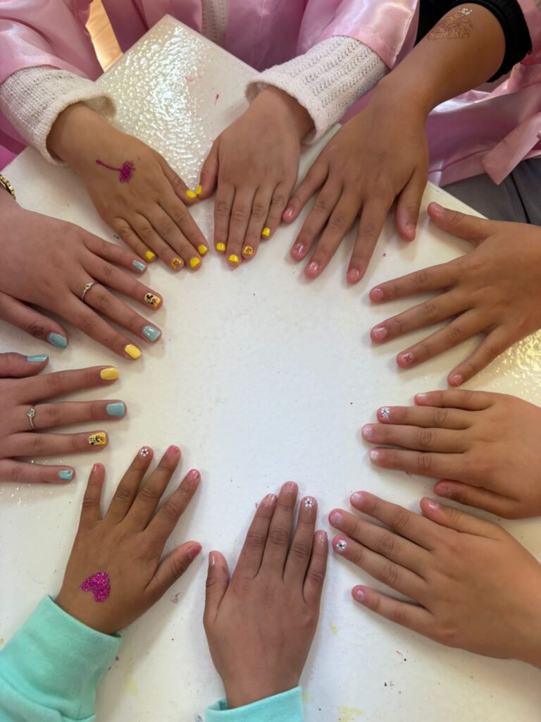 Group of hands with colorful nail art resting on a white table (yellow, turquoise nails with small designs).